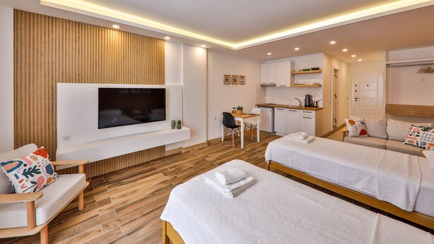 A modern hotel room with a flat-screen TV, two beds, a sofa, a dining area, and a kitchenette.