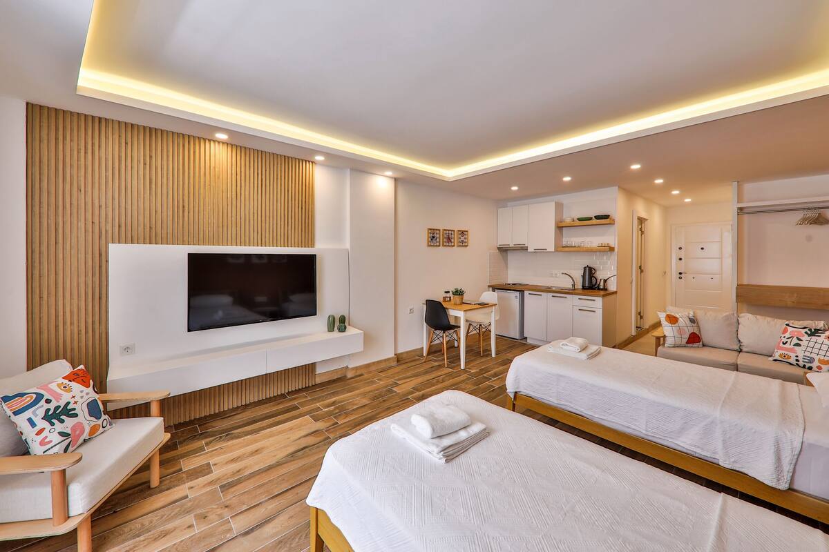 A modern hotel room with a flat-screen TV, two beds, a sofa, a dining area, and a kitchenette.