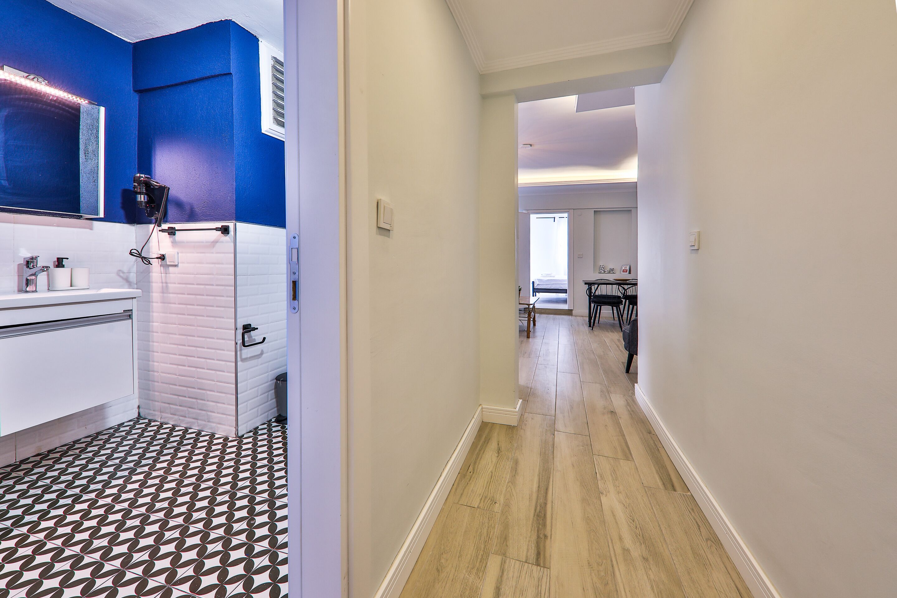 A hallway with a bathroom on the left featuring a white sink and a mirror. The floor has a black and white geometric pattern. The hallway has a wooden floor and white walls.