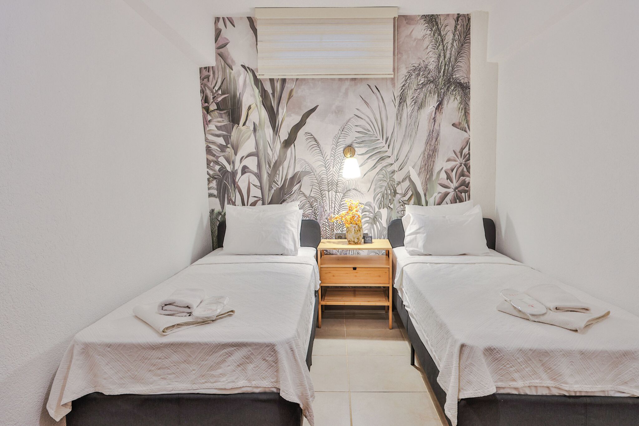 A twin room with two beds, a nightstand with a lamp, and a tropical-themed wall mural.