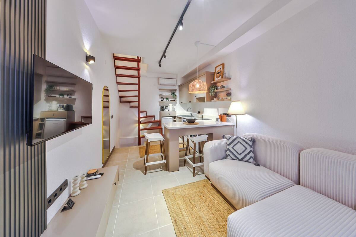 A modern kitchen with a central island, a dining area with bar stools, a staircase, and a living area with a sofa and a TV.
