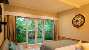 Un dormitorio con un amplio ventanal corredera que da a un jardín, una cama con ropa de cama blanca, un sofá con cojines verdes y una lámpara de pared decorativa.