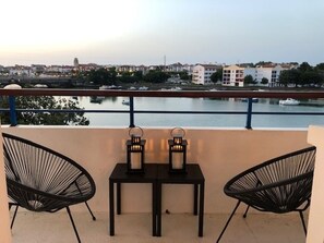 Ein Balkon mit zwei schwarzen Stühlen, einem kleinen Tisch und zwei Laternen. Im Hintergrund ist ein Gewässer und Gebäude zu sehen.