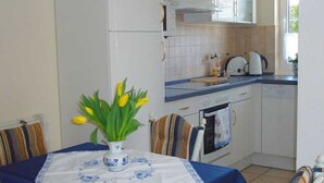 A kitchen with white cabinets, a stove, and a sink. There is a table with a blue tablecloth and a vase with yellow flowers. - Friesenhus - Ferienwohnung (Kühlungsborn)