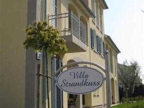 Ein Schild für "Villa Strandkuss" im klassischen Stil, mit einem Balkon und einem gepflegten Garten.