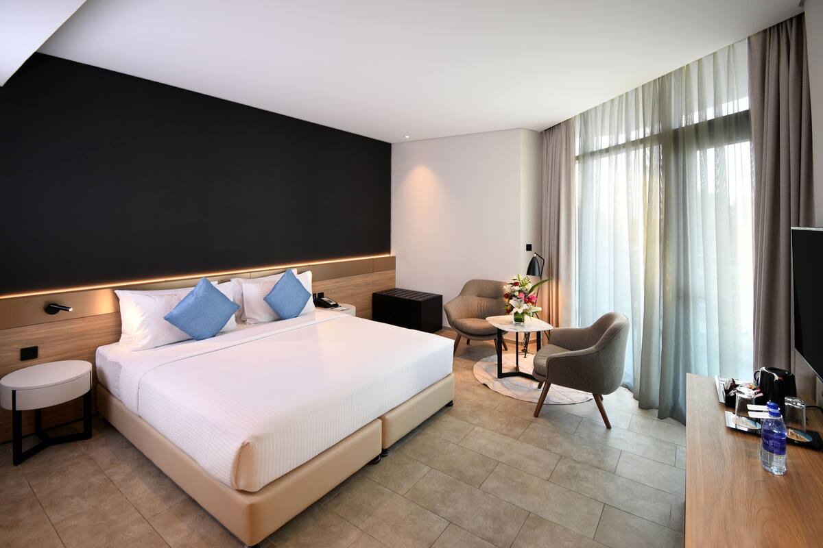 A hotel room with a large bed, bedside tables, a desk, a chair, a TV, and a window with curtains.