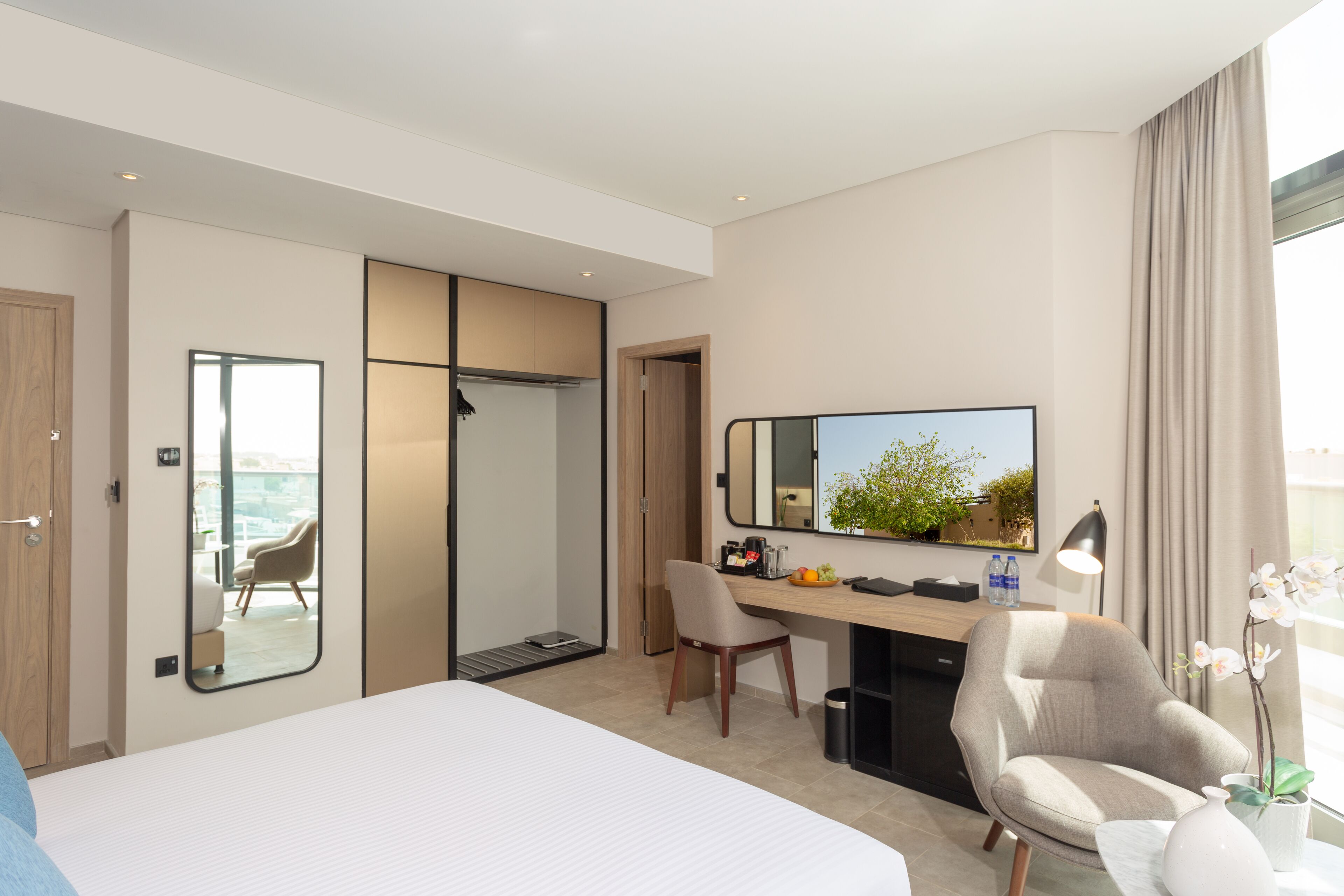 Una habitación de hotel moderna con una cama grande, un escritorio con una silla, un espejo y vistas al exterior.