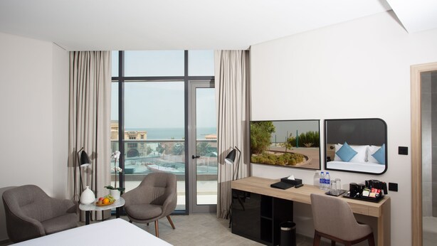 A modern hotel room with a large bed, two armchairs, a desk with a computer, a coffee maker, and a view of the city and ocean. - Beach Walk Hotel (Dubai)