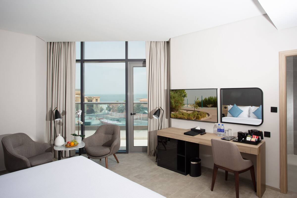 A modern hotel room with a large bed, two armchairs, a desk with a computer, a coffee maker, and a view of the city and ocean.