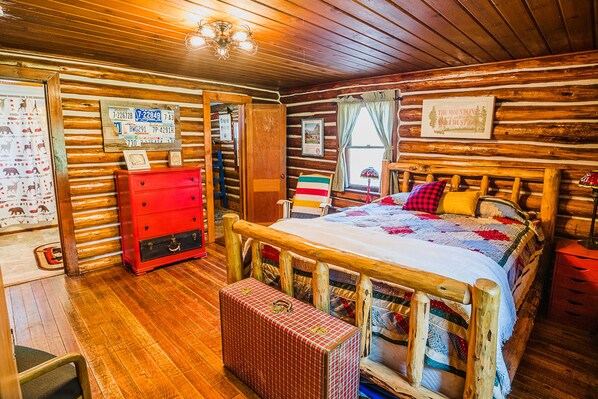 A log cabin-style room with a wooden bed, a red dresser, and a quilted blanket. - Authentic Vintage Cabin 9 Miles from Glacier (Martin City)