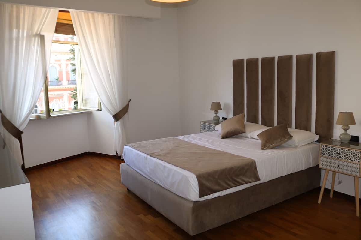 A bedroom with a large bed, two bedside tables, a window with curtains, and a view of buildings outside.