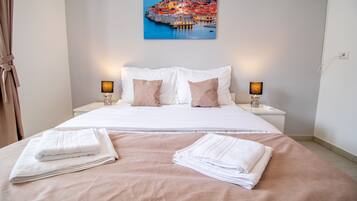 A neatly made bed with white and beige linens, two bedside lamps, and a painting of a coastal town above the bed.