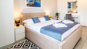 A modern hotel room with a large bed, a bedside table with a lamp, a desk with a chair, a mirror, and a painting of a coastal scene.