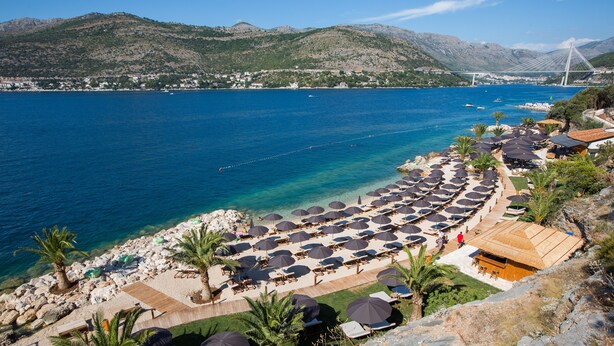 A beachside resort with sun loungers and umbrellas, a wooden deck, and a view of the sea and mountains.