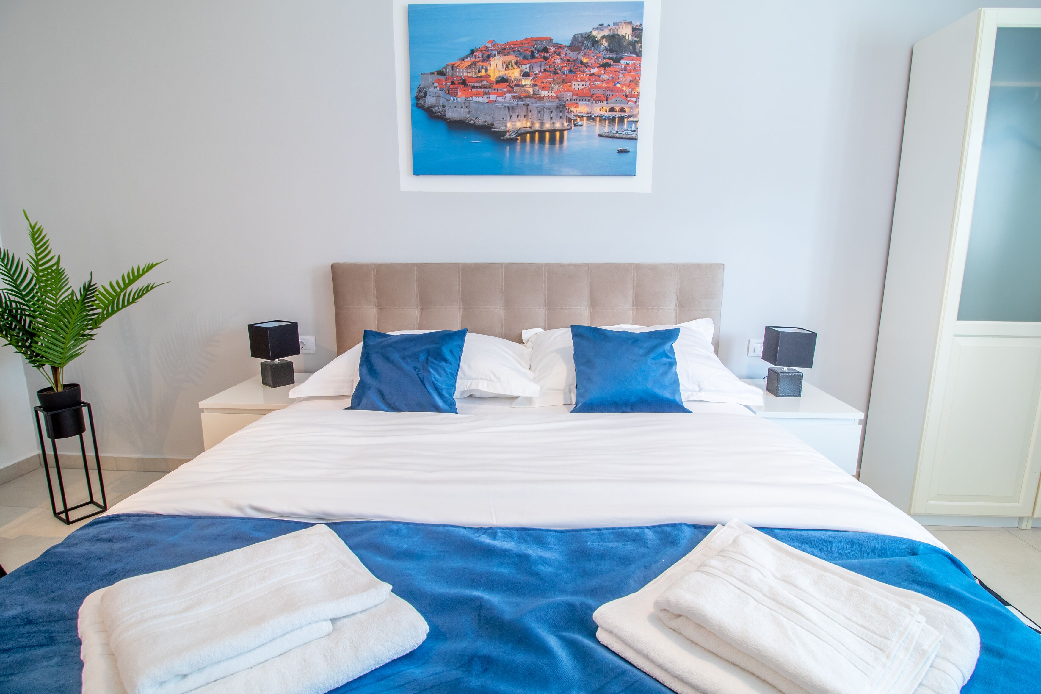 Uma cama bem arrumada com roupa de cama branca e azul, um vaso de planta e um quadro com uma paisagem costeira na parede.