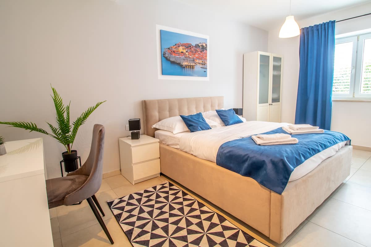 Ein modernes Schlafzimmer mit einem großen Bett, einem Nachttisch, einem Sessel, einer Topfpflanze und einem geometrischen Teppich.