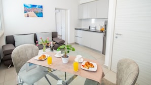 A modern living room with a glass dining table, two sofas, a coffee table with breakfast items, and a kitchen in the background.