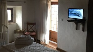 A room with a wooden ceiling, a television mounted on the wall, a chair, a bed with a white blanket, and a window with curtains.