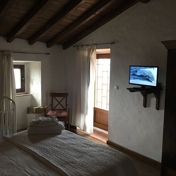 A room with a wooden ceiling, a television mounted on the wall, a chair, a bed with a white blanket, and a window with curtains.