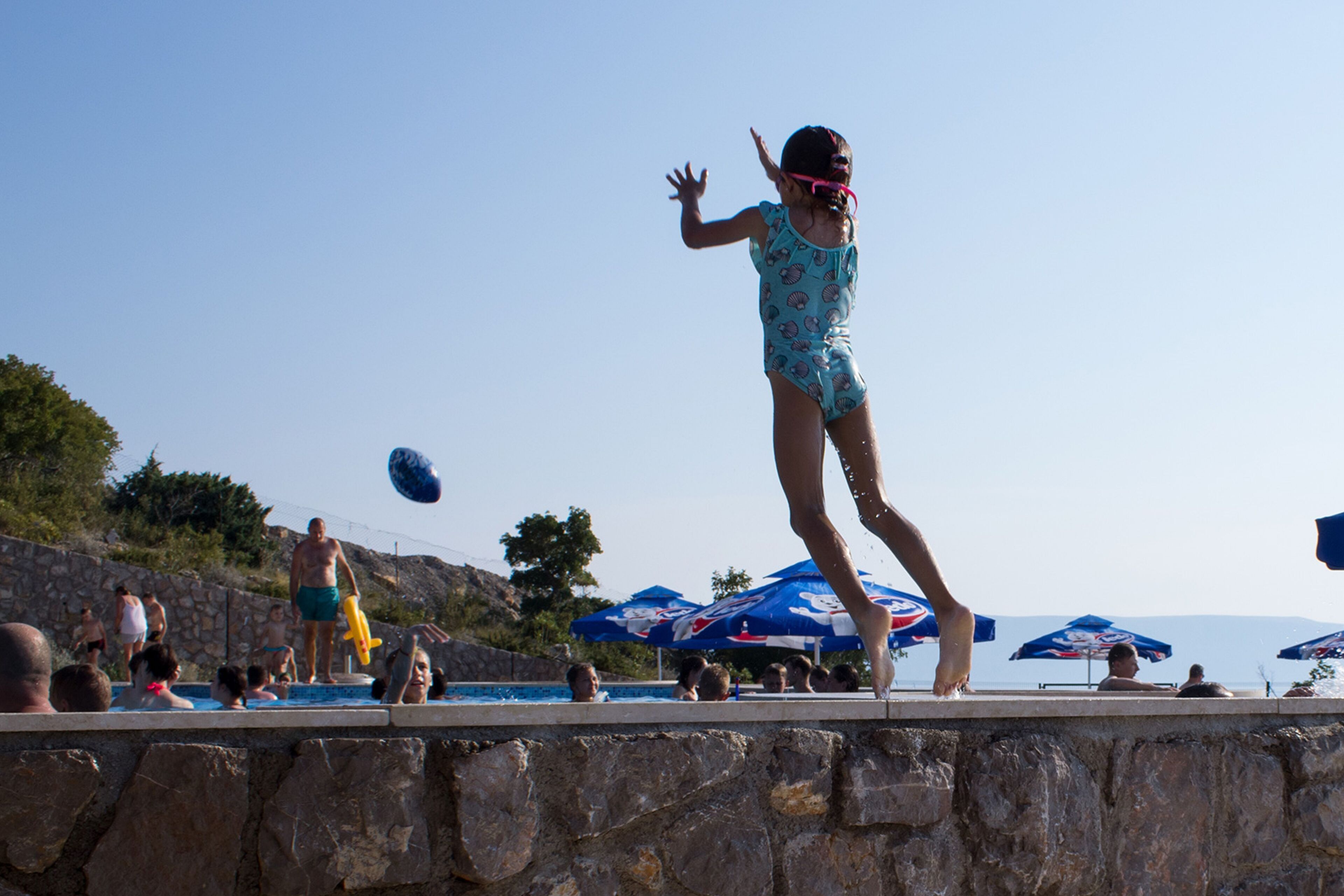 Zona de piscina con paredes de piedra, gente nadando y sombrillas al fondo.