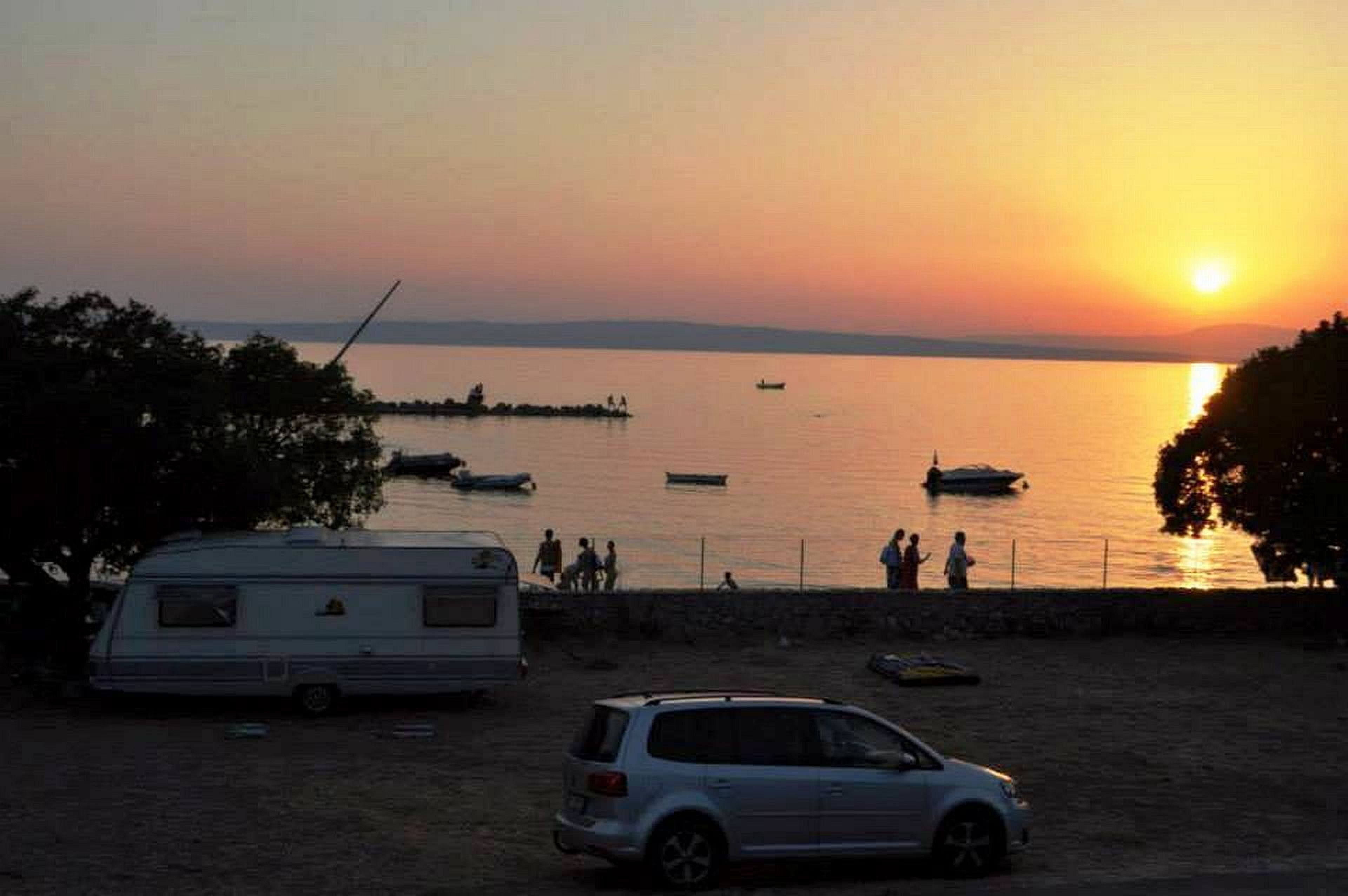 Une scène de plage avec un camping-car et une voiture garés sur le sable, un coucher de soleil en arrière-plan et plusieurs bateaux sur l’eau.