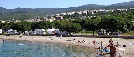 A beach with clear water, people sunbathing, and a hillside with houses in the background.