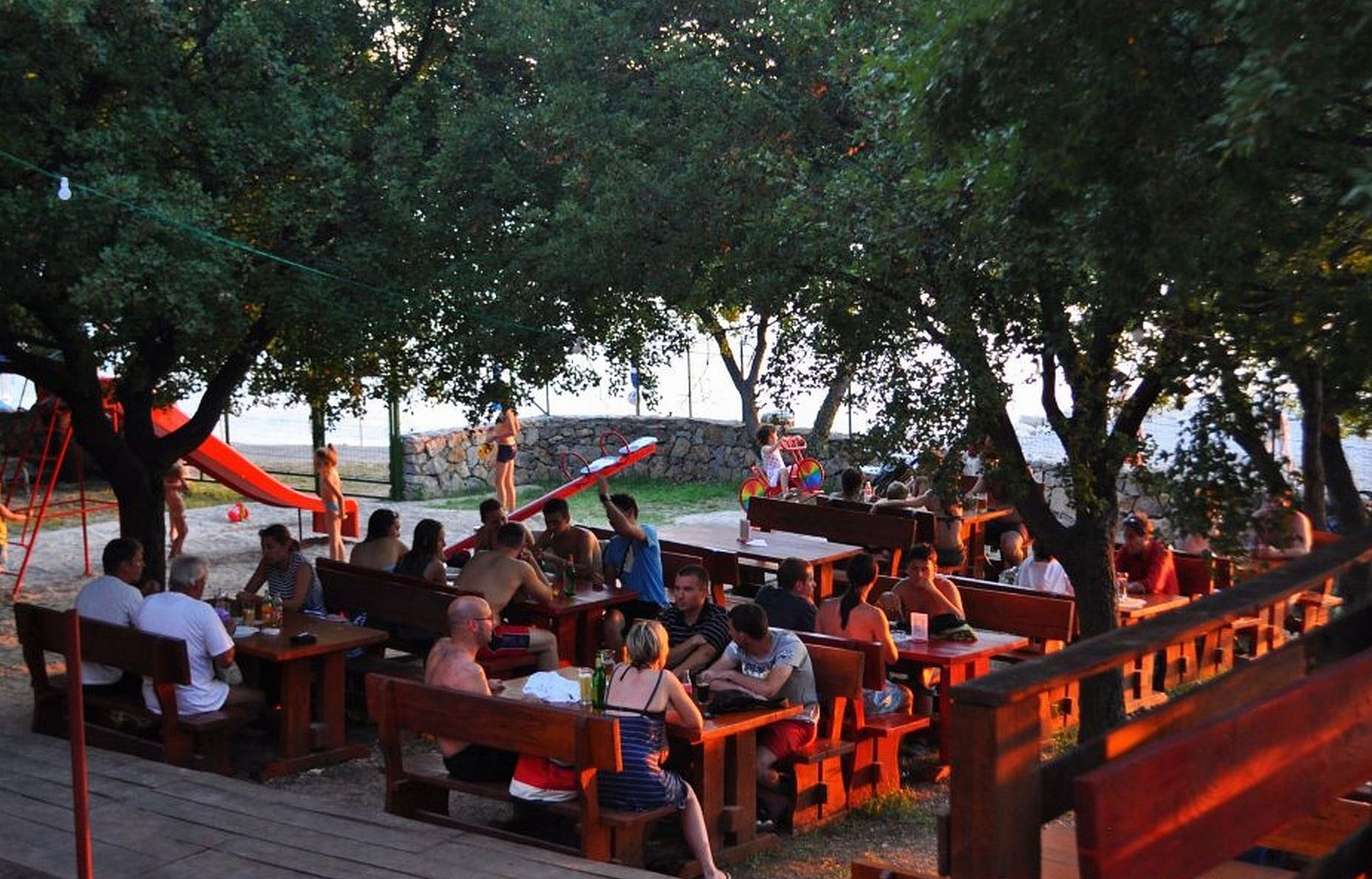 An outdoor dining area with wooden tables and benches, surrounded by trees and a stone wall.