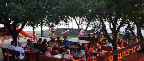 An outdoor dining area with wooden tables and benches, surrounded by trees and a stone wall.