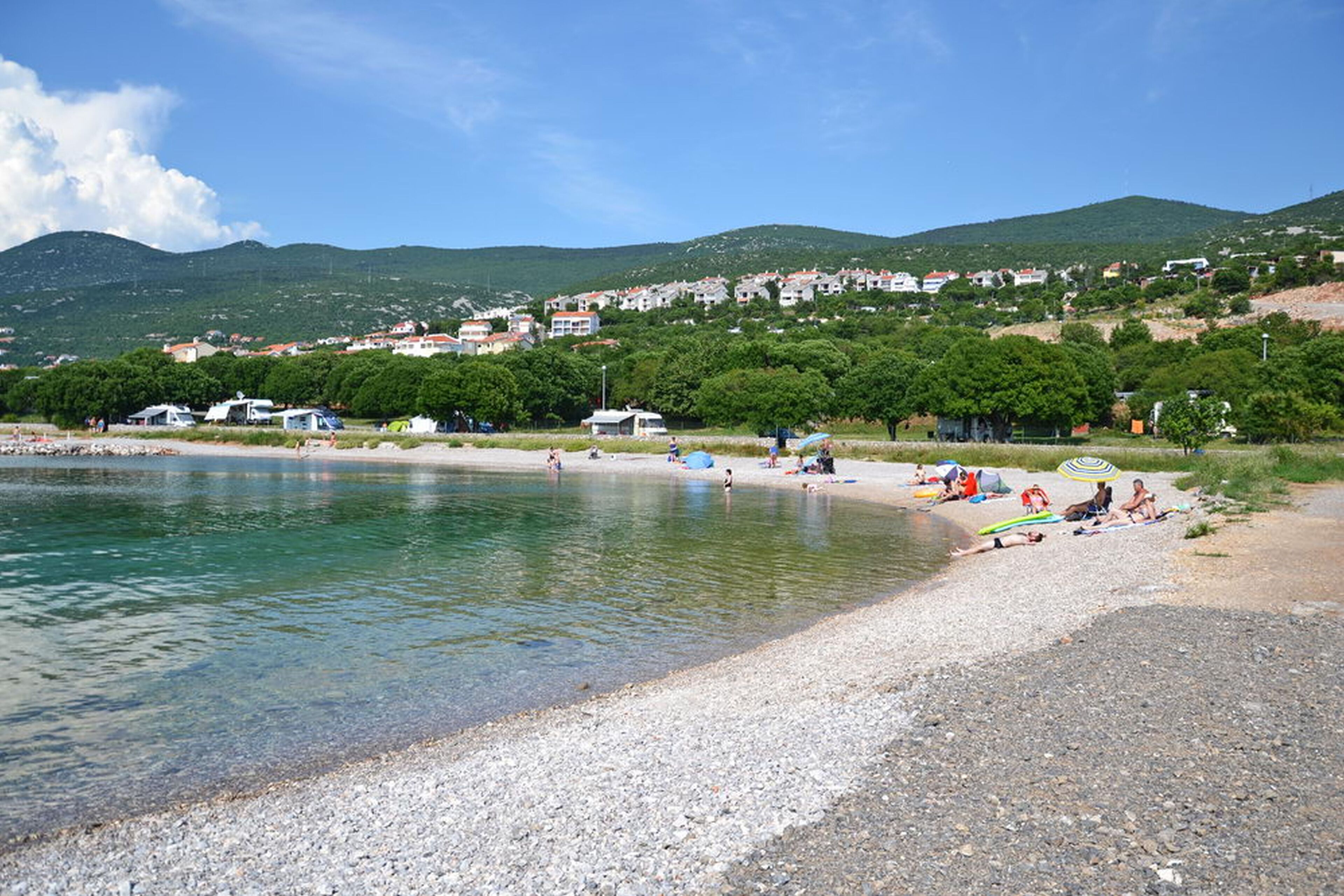 A beach with clear water, pebbles, and people relaxing. There are houses and trees in the background.