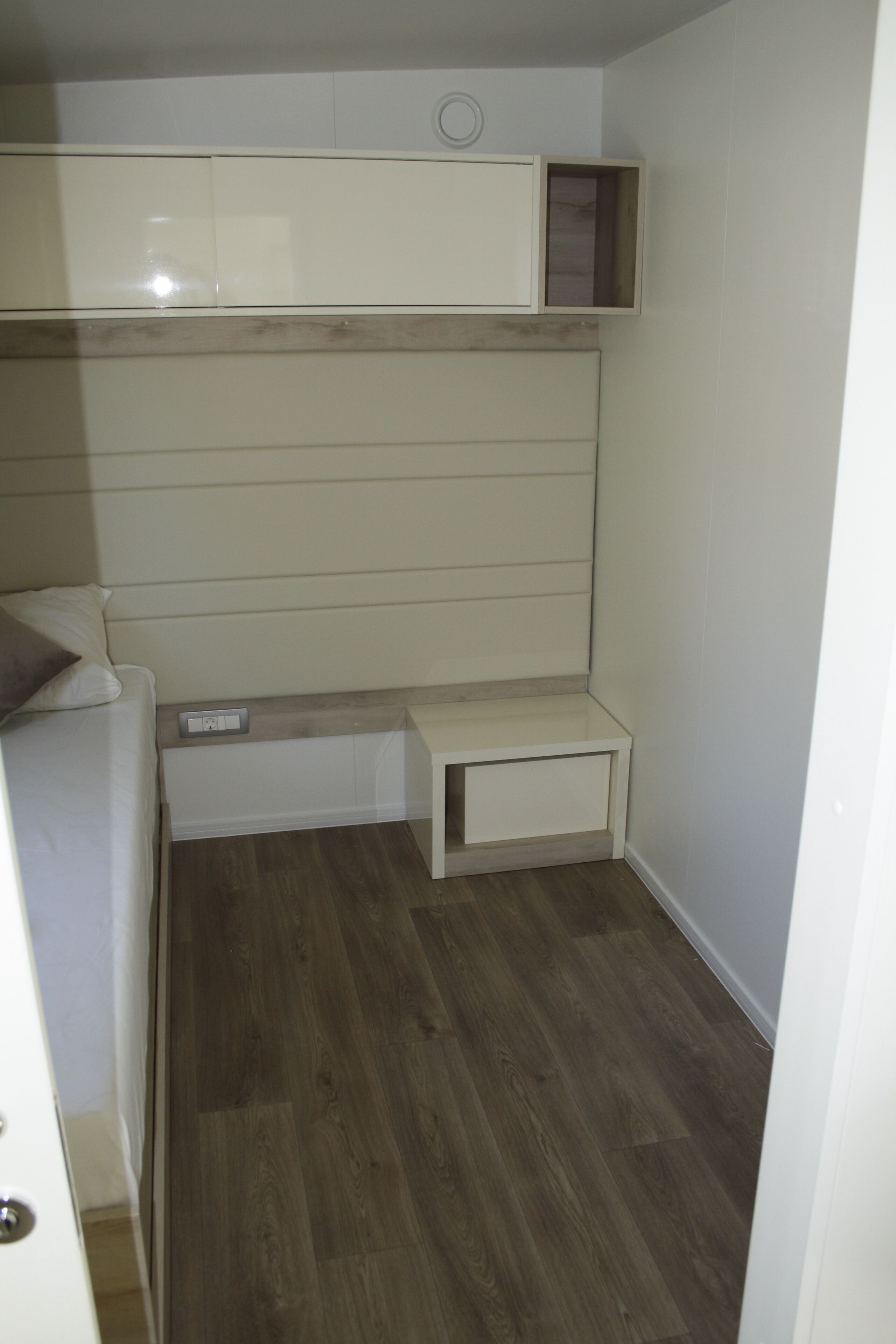 Een kleine kamer met een bed, nachtkastje en een inbouwplank boven het bed.