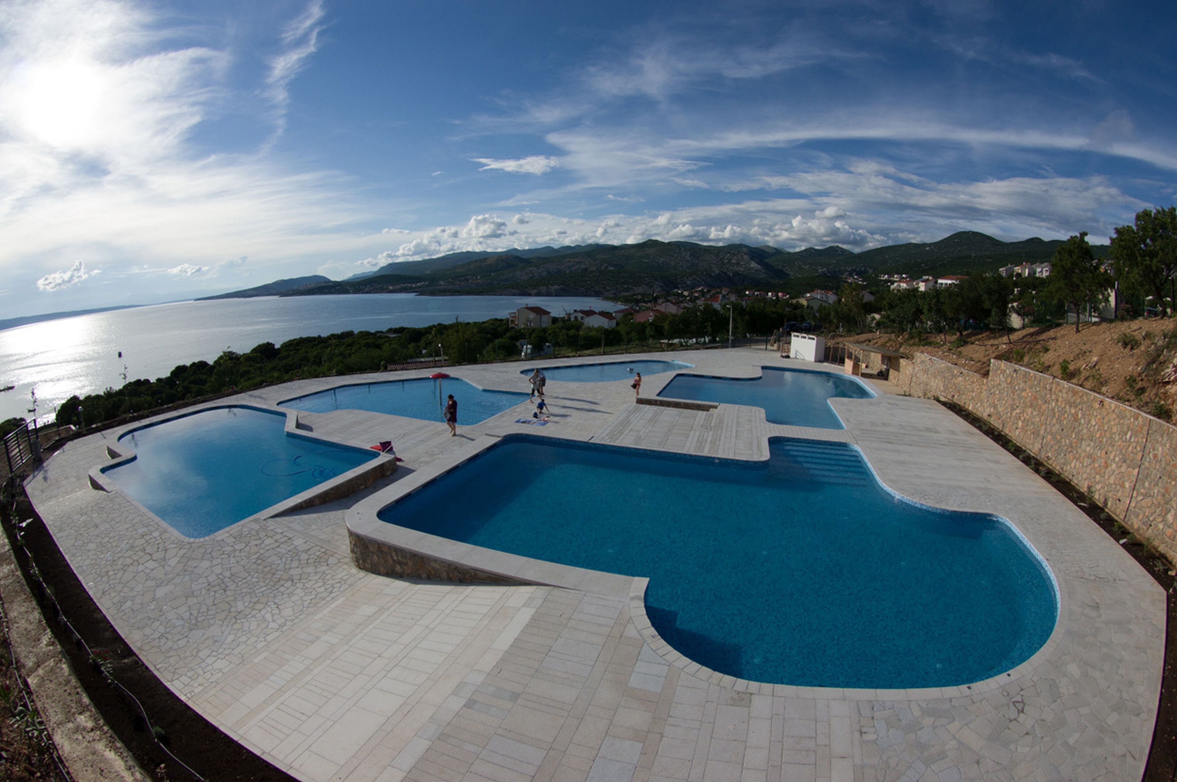 A large, uniquely shaped swimming pool with multiple sections, surrounded by a stone deck and overlooking a scenic body of water and distant hills.