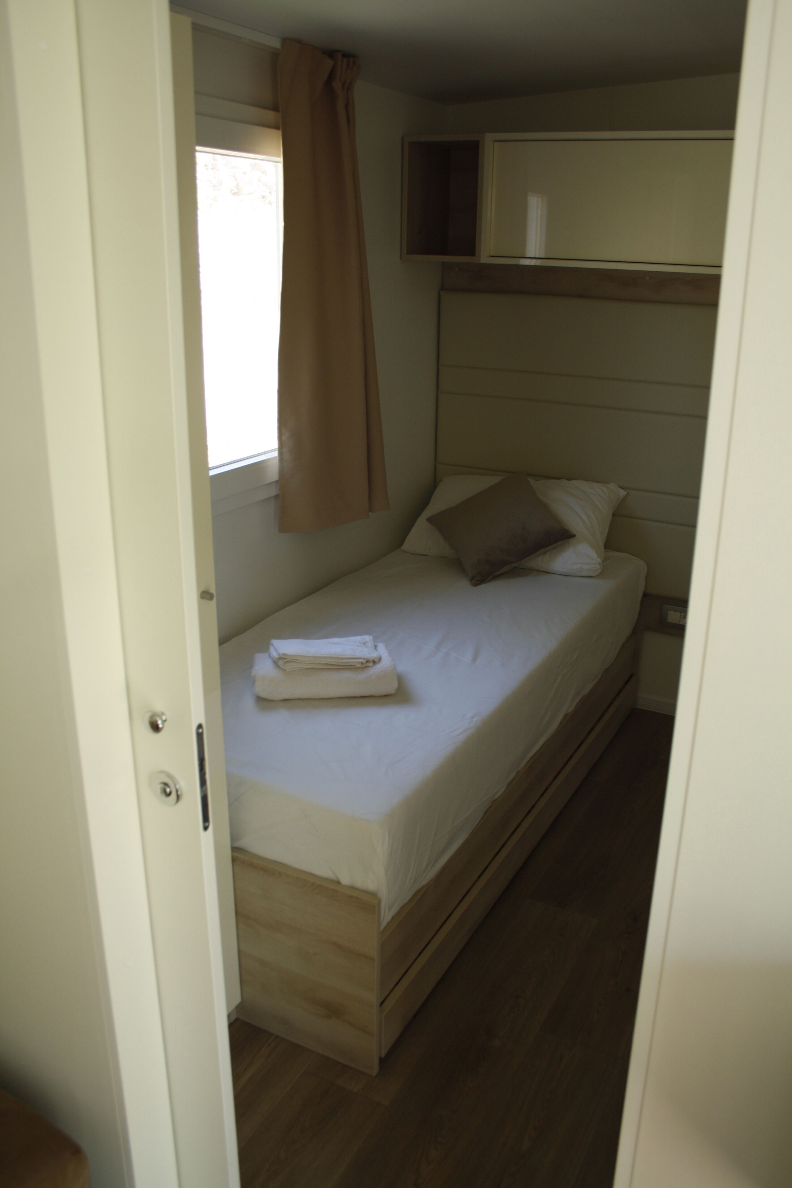 A small, single-bed room with a wooden bed frame, a window with a curtain, and a bedside table with a lamp.
