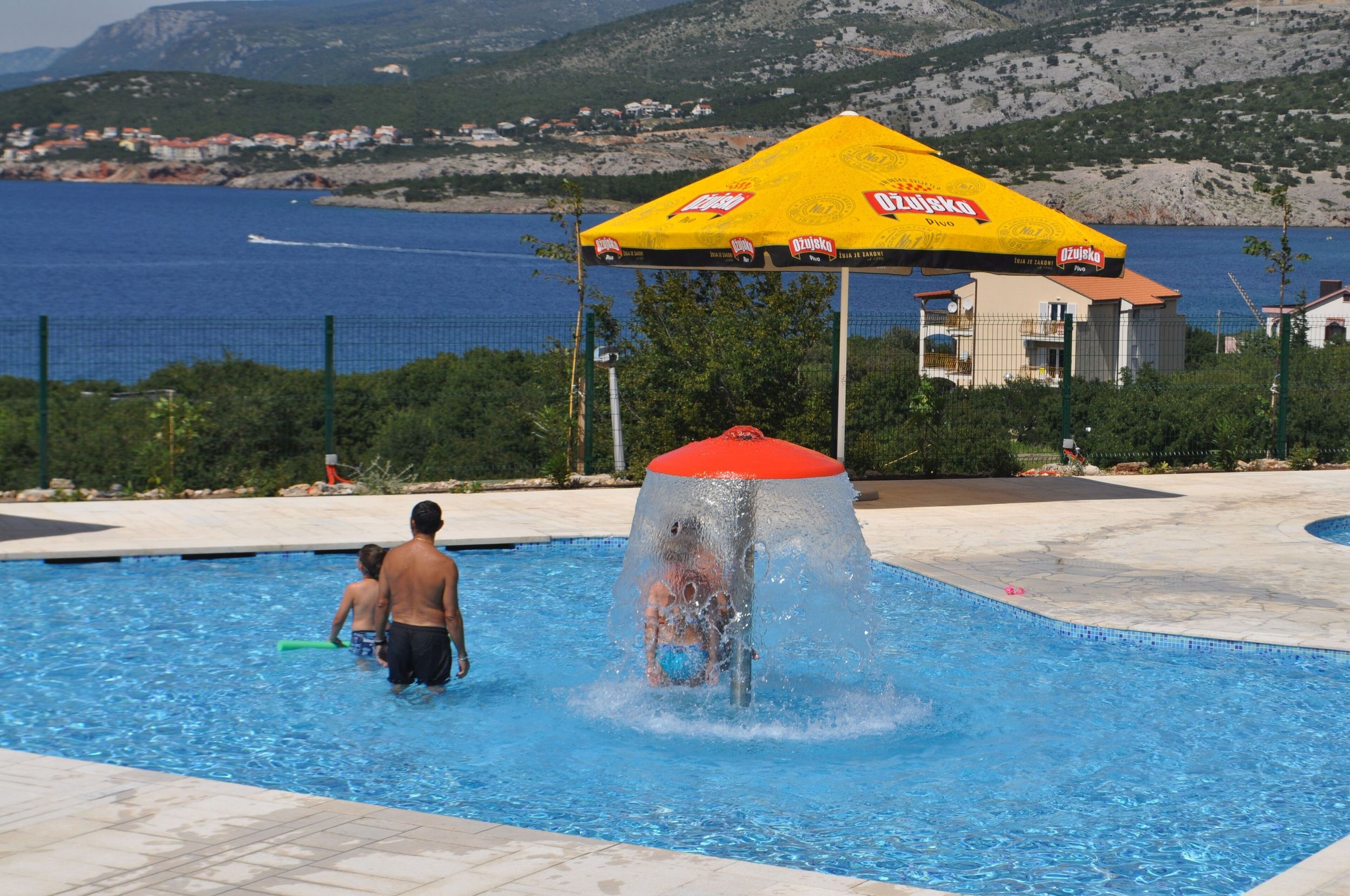 A swimming pool with a water fountain feature, surrounded by a fence and a yellow umbrella.