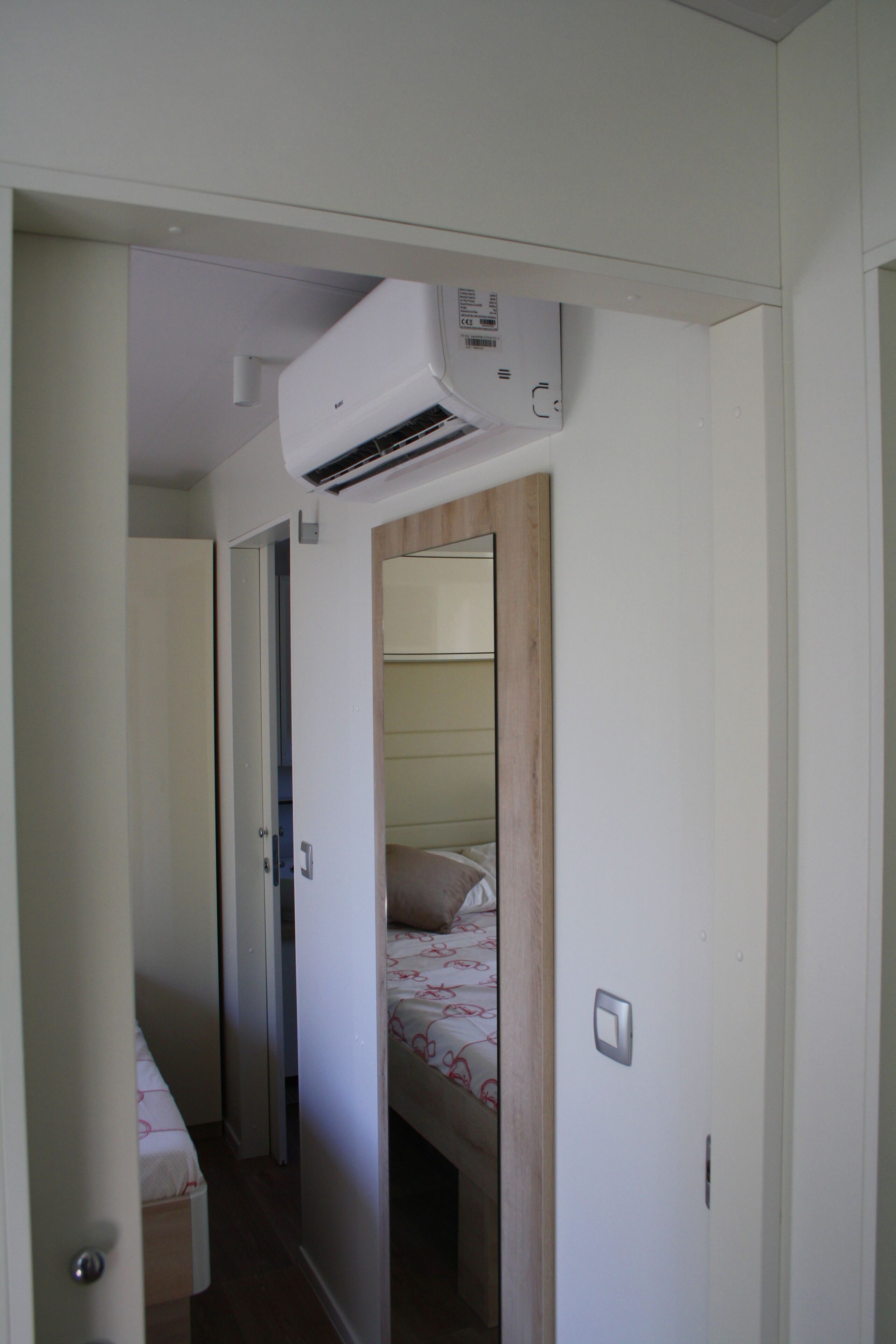 Ein schmaler Flur mit einem Bett am Ende, einer Klimaanlage darüber und einem spiegelnden Wandbild mit Holzrahmen.