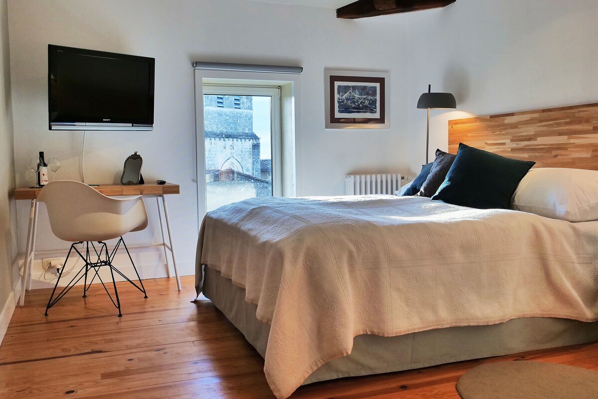 Ein Schlafzimmer mit Bett, Schreibtisch, Stuhl, Fernseher, Fenster mit Blick auf ein Gebäude und Heizkörper.