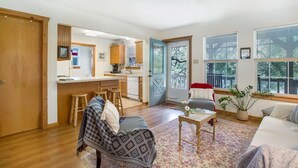 A cozy living room with a sofa, a coffee table, and a chair. There is a kitchen area with a counter and stools. A door leads to a balcony with a view of trees. - Creekside Country Guesthouse | 2 Bedroom plus loft | Beautiful Country Setting (Fredericksburg)