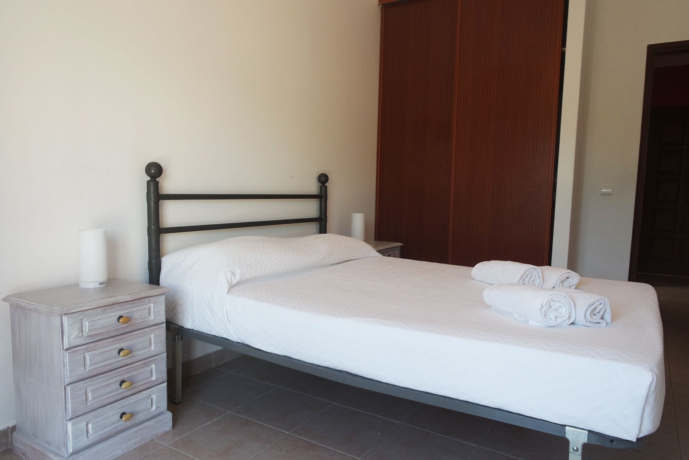 A neatly made bed with white linens and rolled towels, a wooden nightstand with drawers, and a wooden wardrobe in the background.