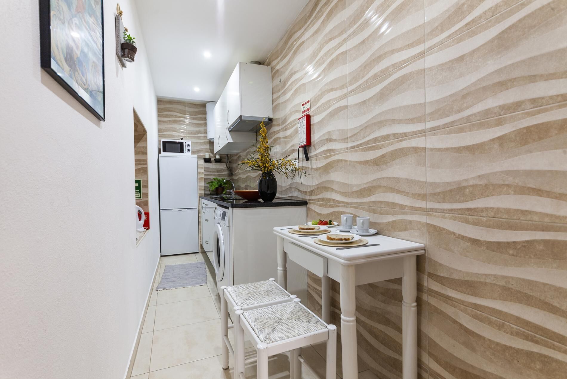 A narrow kitchen with a microwave, refrigerator, and sink. There is a small table with plates and cups, and a bench. The walls have a wavy pattern.