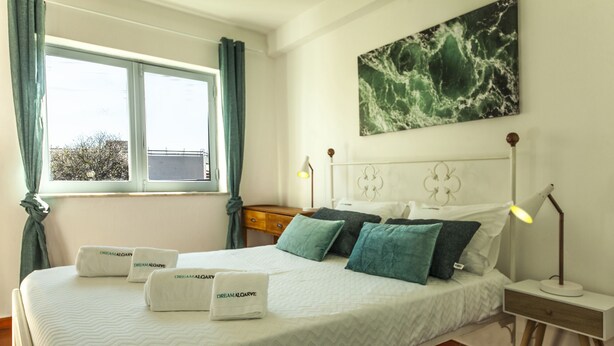 Una cama bien hecha con una colcha blanca y varios almohadones, incluyendo unos color turquesa y blanco, y dos toallas con el texto 'Dream Alomarve'.