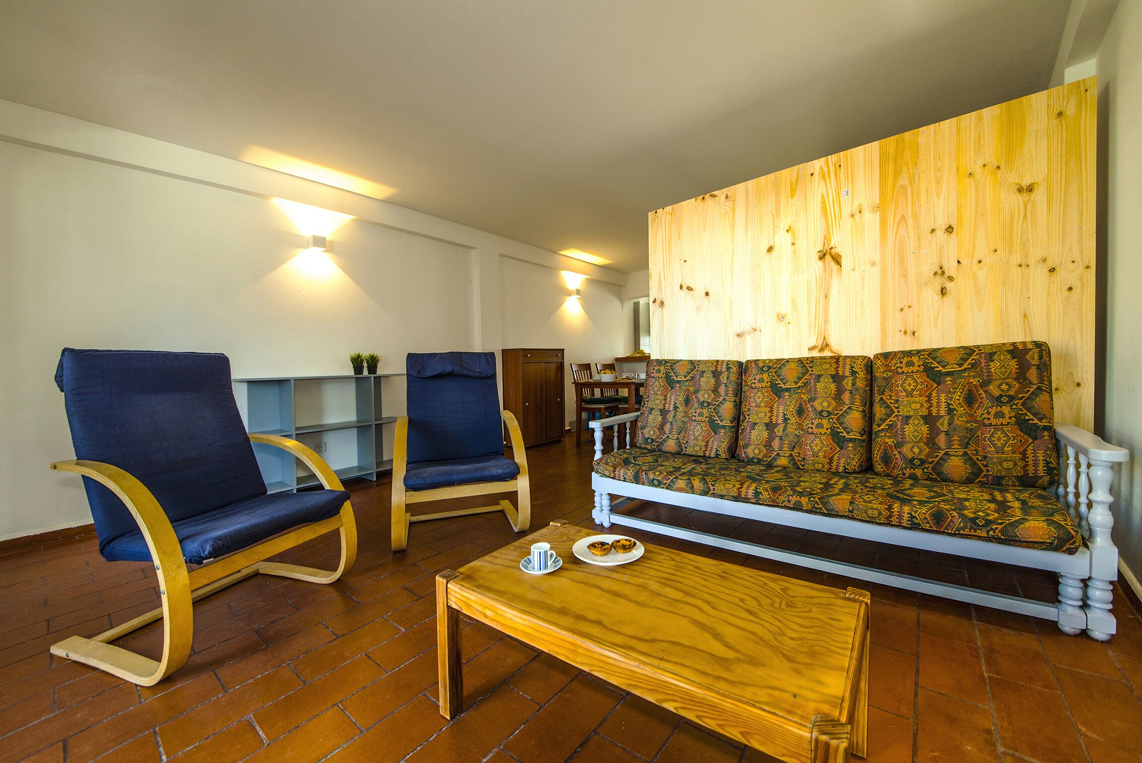 A living room with a patterned sofa, two blue armchairs, a wooden coffee table, and a wall with a wooden panel design.