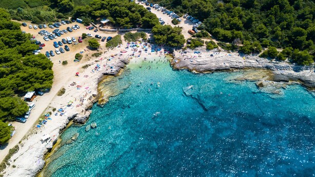 Vista aérea de una zona costera con playa, aguas azules cristalinas y un estacionamiento con numerosos vehículos.