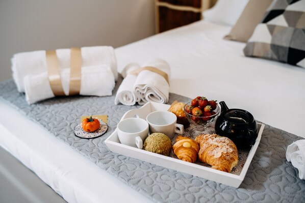 Una cama bien hecha con sábanas blancas y una bandeja de desayuno que incluye cruasanes, fresas y una naranja pequeña.
