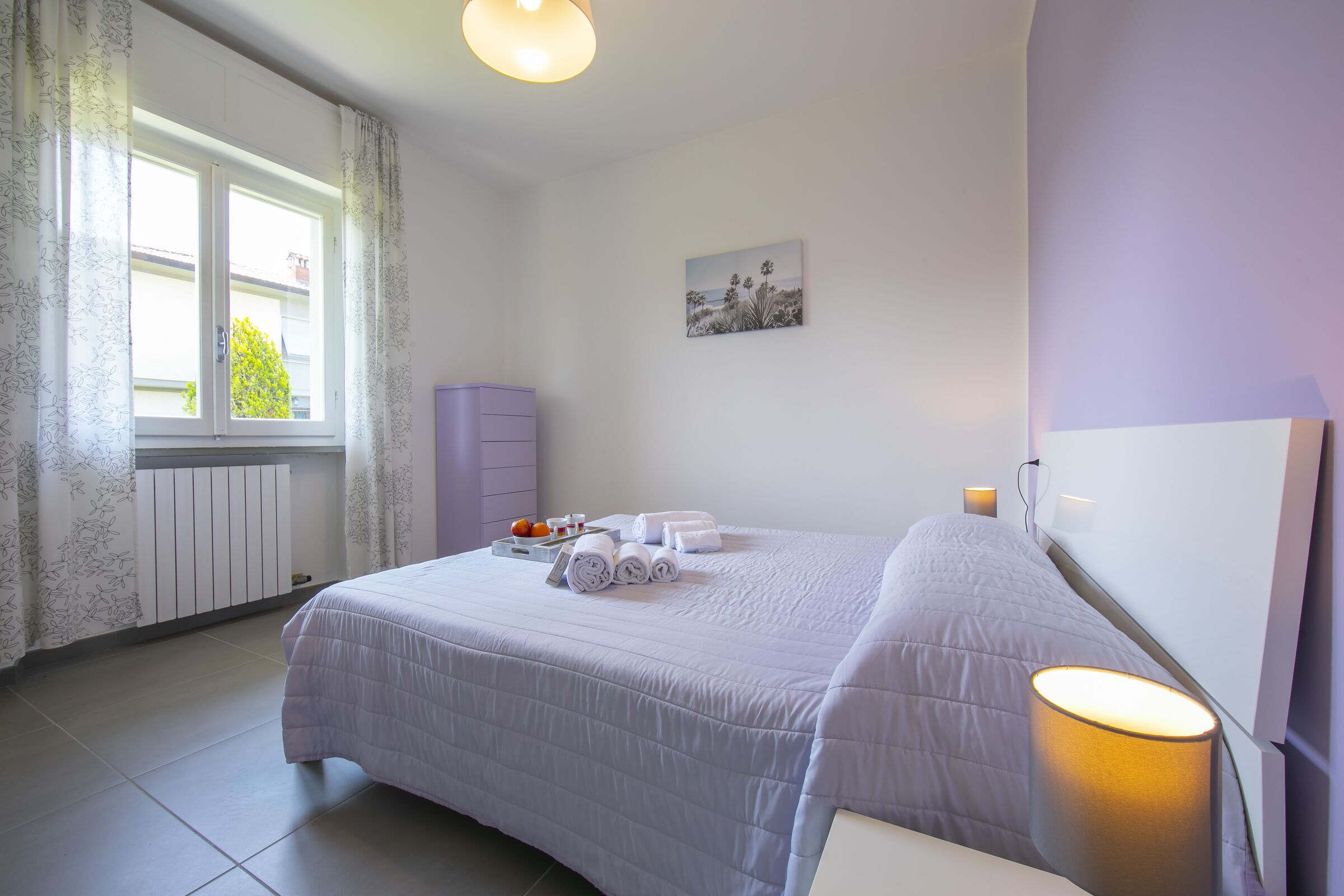 A bedroom with a bed, a bedside lamp, a radiator, a window with curtains, and a wall-mounted picture.
