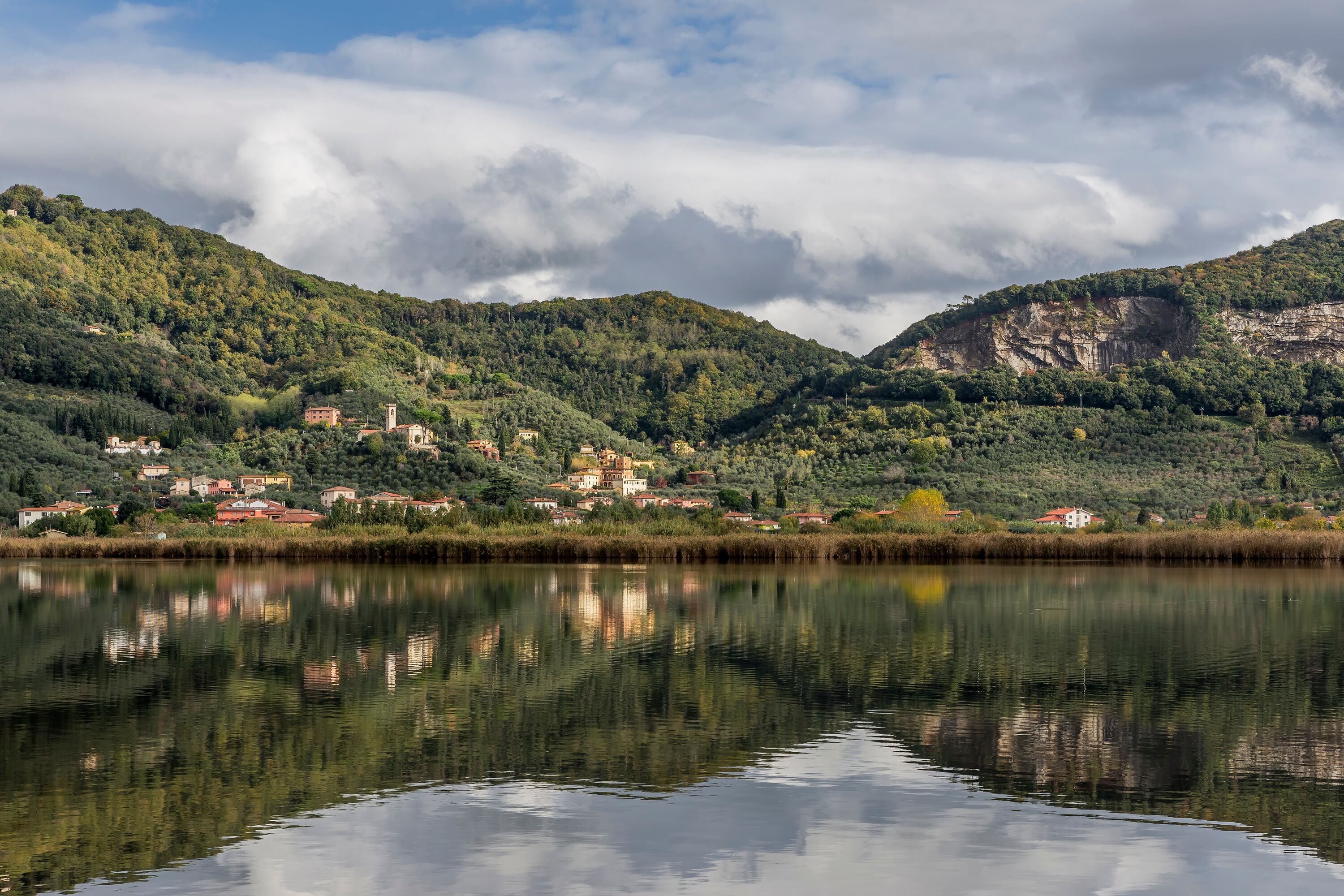 Vista de un lago con edificios en una colina, agua tranquila que refleja las construcciones y un cielo nublado.