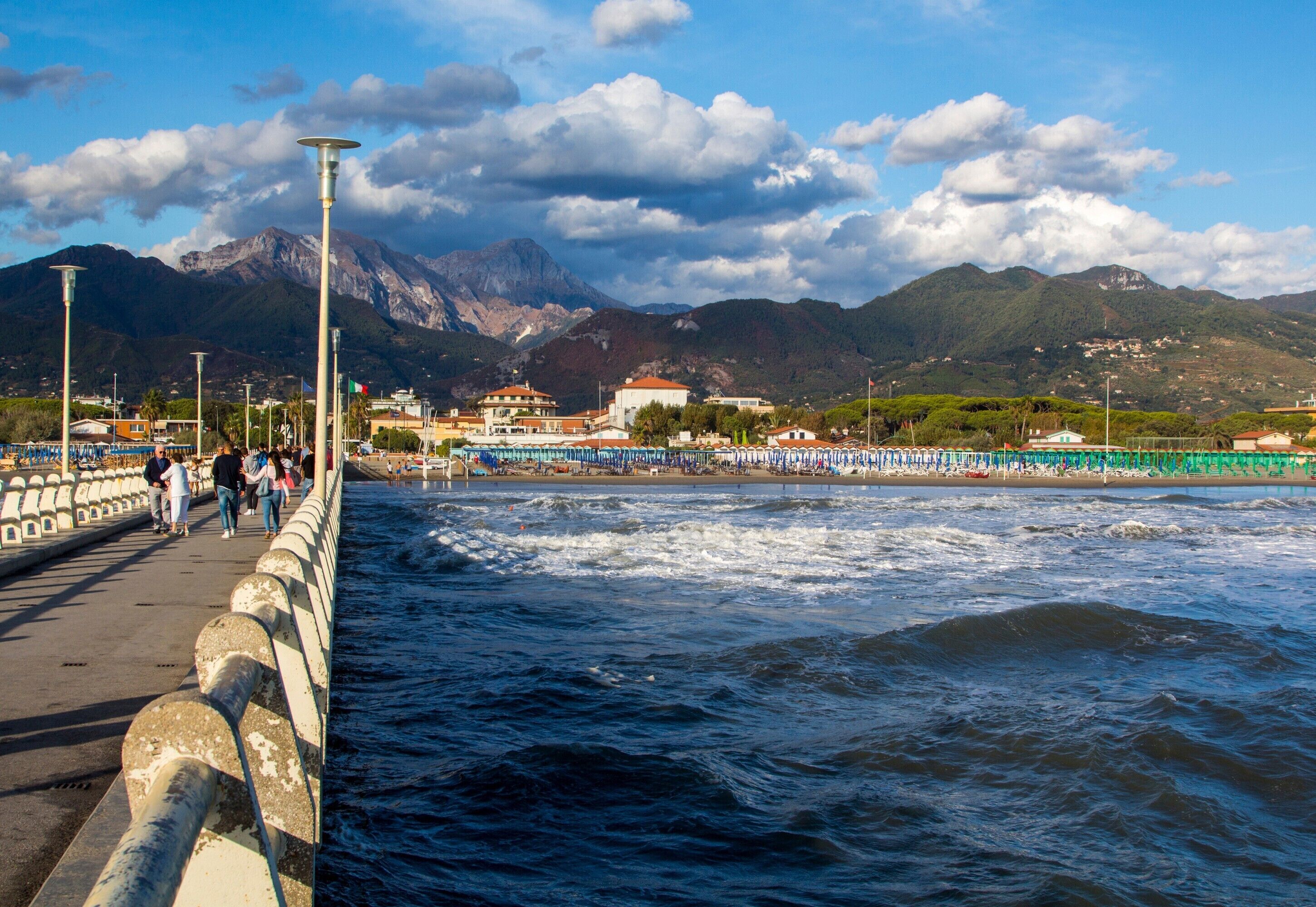 A coastal promenade with a view of mountains, a beach, and a pier with people walking.