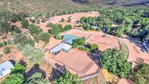 An aerial view of a rural property with a fenced horse riding arena, several buildings, and surrounding forested hills.
