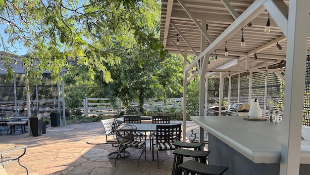 An outdoor patio area with stone flooring, metal tables and chairs, a bar counter, and a pergola with hanging lights.
