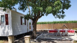Una casa móvil con un árbol al frente, un área cercada con una mesa y sillas, y un campo de maíz al fondo.