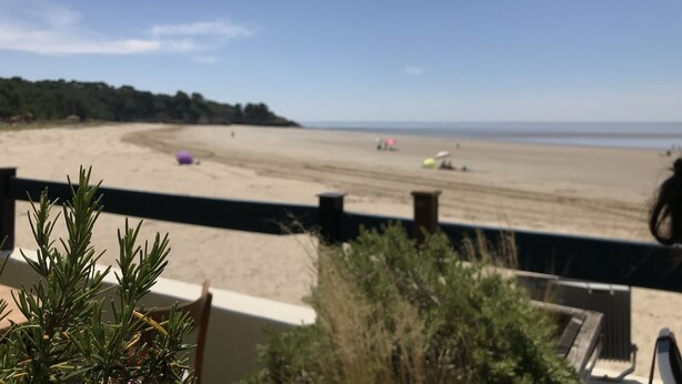 Vista a la playa desde un balcón con barandilla, una silla y algo de vegetación en primer plano.