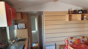 Una cocina compacta con microondas, tetera y lavamanos. Hay una mesita con una silla y un estante encima.